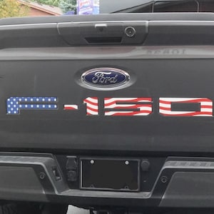May include: A dark gray Ford F-150 truck tailgate featuring a blue and white star-patterned decal on the letter F and red and white striped decals on the numbers 150. A small black American flag decal is positioned to the left of the Ford emblem.