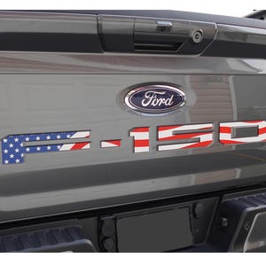 May include: A gray Ford F-150 truck tailgate featuring custom emblem inserts. The letters and numbers display a patriotic American flag design with white stars on a blue field on the F, and red and white horizontal stripes across the 150.