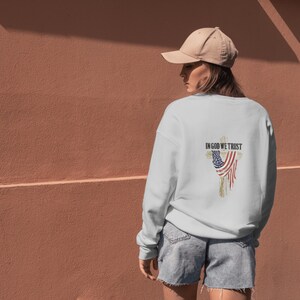 Puede incluir: Sudadera blanca con un dise&ntilde;o desgastado de la bandera estadounidense y el texto "IN GOD WE TRUST" en la espalda. La sudadera se combina con pantalones cortos de mezclilla y una gorra de b&eacute;isbol beige.