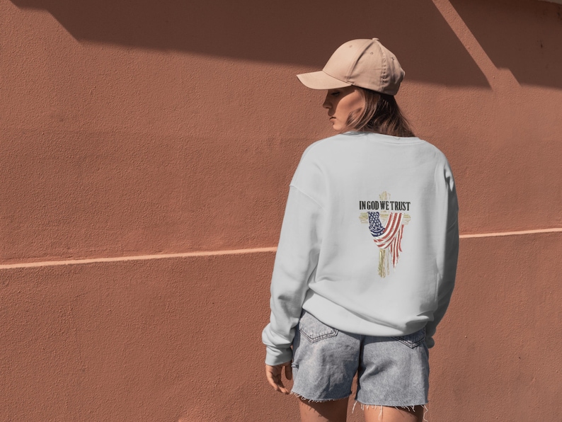 Puede incluir: Sudadera blanca con un dise&ntilde;o desgastado de la bandera estadounidense y el texto "IN GOD WE TRUST" en la espalda. La sudadera se combina con pantalones cortos de mezclilla y una gorra de b&eacute;isbol beige.