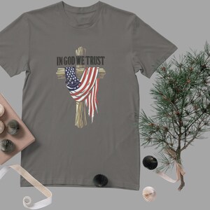 Puede incluir: Camiseta gris con un dise&ntilde;o de cruz con una bandera estadounidense. El texto "IN GOD WE TRUST" est&aacute; encima de la cruz. La camiseta es de cuello redondo.