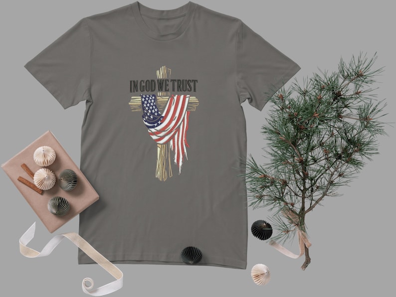 Puede incluir: Camiseta gris con un dise&ntilde;o de cruz con una bandera estadounidense. El texto "IN GOD WE TRUST" est&aacute; encima de la cruz. La camiseta es de cuello redondo.