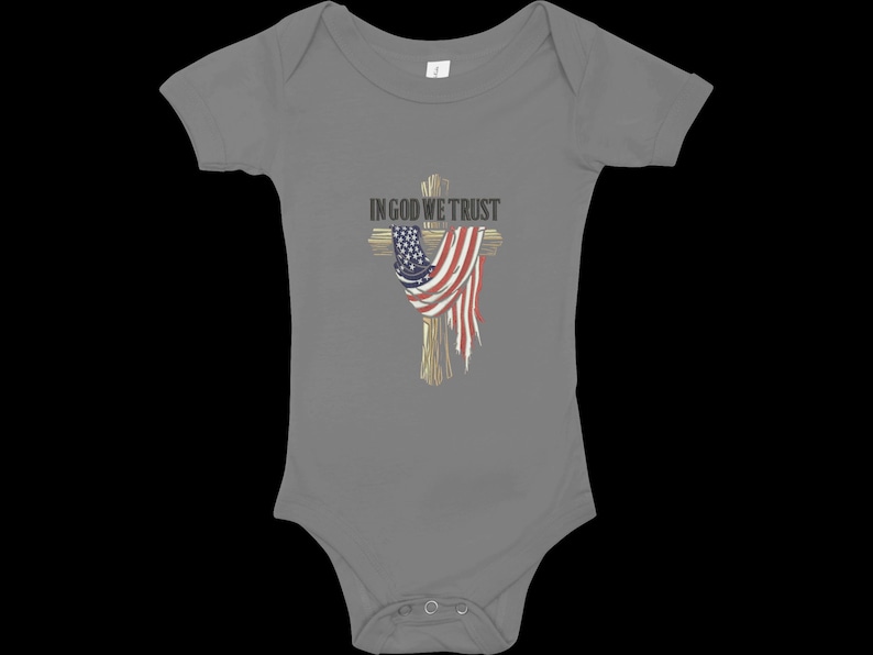 Puede incluir: Body de beb&eacute; gris con un dise&ntilde;o de cruz de madera adornada con una bandera americana. El texto "IN GOD WE TRUST" est&aacute; sobre la cruz. El body tiene mangas cortas y cierres de presi&oacute;n.