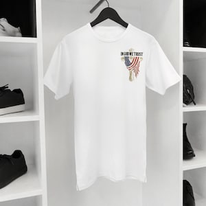 Puede incluir: Camiseta blanca con un peque&ntilde;o gr&aacute;fico en la parte superior izquierda del pecho. El gr&aacute;fico presenta el texto "IN GOD WE TRUST" sobre un dise&ntilde;o estilizado de la bandera estadounidense. La camiseta est&aacute; colgada en una percha negra.