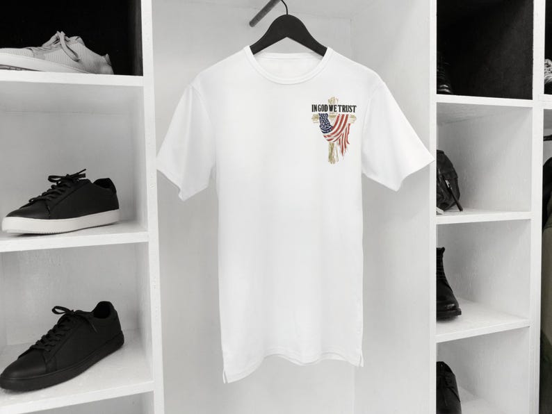 Puede incluir: Camiseta blanca con un peque&ntilde;o gr&aacute;fico en la parte superior izquierda del pecho. El gr&aacute;fico presenta el texto "IN GOD WE TRUST" sobre un dise&ntilde;o estilizado de la bandera estadounidense. La camiseta est&aacute; colgada en una percha negra.