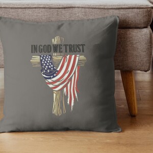 Puede incluir: Coj&iacute;n decorativo gris con una cruz de madera adornada con una bandera estadounidense desgastada. Las palabras "IN GOD WE TRUST" est&aacute;n impresas en negrita sobre la cruz. El coj&iacute;n est&aacute; sobre un suelo de madera.