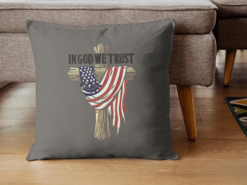 Puede incluir: Coj&iacute;n decorativo gris con una cruz de madera adornada con una bandera estadounidense desgastada. Las palabras "IN GOD WE TRUST" est&aacute;n impresas en negrita sobre la cruz. El coj&iacute;n est&aacute; sobre un suelo de madera.