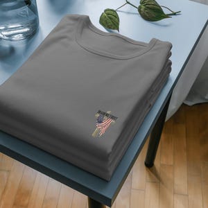 Puede incluir: Una pila de tres camisetas grises con cuello redondo. Cada camiseta tiene un peque&ntilde;o gr&aacute;fico de una cruz con un dise&ntilde;o de la bandera estadounidense y las palabras "IN GOD WE TRUST". Las camisetas est&aacute;n cuidadosamente dobladas sobre una mesa azul claro.