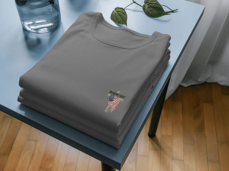 Puede incluir: Una pila de tres camisetas grises con cuello redondo. Cada camiseta tiene un peque&ntilde;o gr&aacute;fico de una cruz con un dise&ntilde;o de la bandera estadounidense y las palabras "IN GOD WE TRUST". Las camisetas est&aacute;n cuidadosamente dobladas sobre una mesa azul claro.