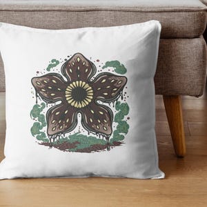 May include: White throw pillow featuring a Demogorgon design. The brown, flower-like creature has a textured pattern and a yellow center. Green and brown accents surround the central image, creating a unique decorative item.