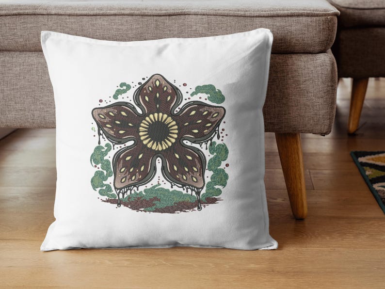 May include: White throw pillow featuring a Demogorgon design. The brown, flower-like creature has a textured pattern and a yellow center. Green and brown accents surround the central image, creating a unique decorative item.