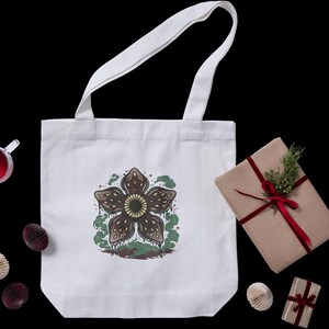 May include: White tote bag featuring a Demogorgon graphic in brown, green, and yellow. The bag has white handles and is on a black background. A wrapped gift with a red ribbon and a sprig of greenery is next to the bag.