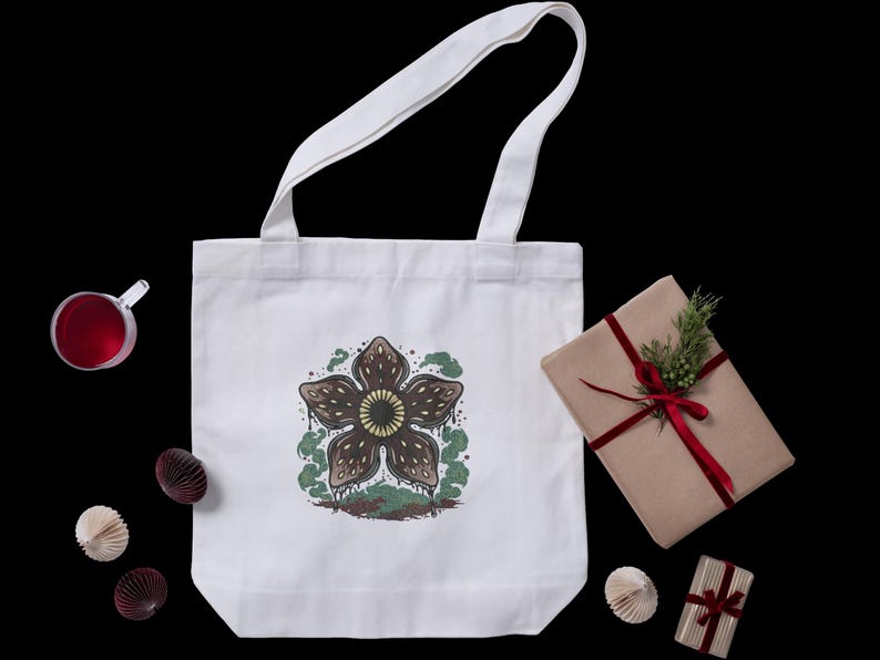 May include: White tote bag featuring a Demogorgon graphic in brown, green, and yellow. The bag has white handles and is on a black background. A wrapped gift with a red ribbon and a sprig of greenery is next to the bag.