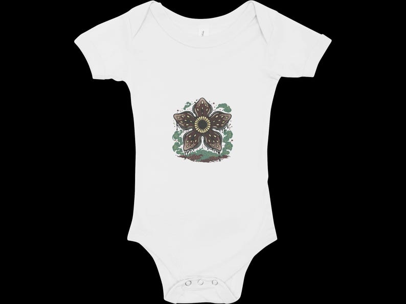 May include: White baby onesie featuring a Demogorgon flower graphic. The graphic is brown, green, and tan. The onesie has short sleeves and snaps at the bottom. A cute and unique baby outfit.