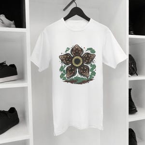 May include: White t-shirt featuring a Demogorgon flower design. The graphic has a brown flower with a yellow center, surrounded by green and brown accents. The shirt is hanging on a black hanger.