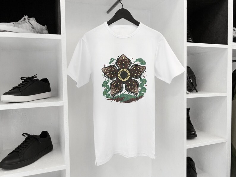 May include: White t-shirt featuring a Demogorgon flower design. The graphic has a brown flower with a yellow center, surrounded by green and brown accents. The shirt is hanging on a black hanger.