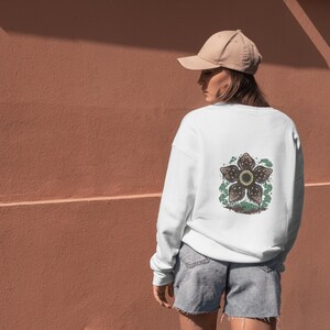 May include: White sweatshirt with a Demogorgon graphic on the back. The graphic is a detailed illustration of the creature's flower-like head, with brown and green colors. The person is wearing a tan baseball cap and denim shorts.