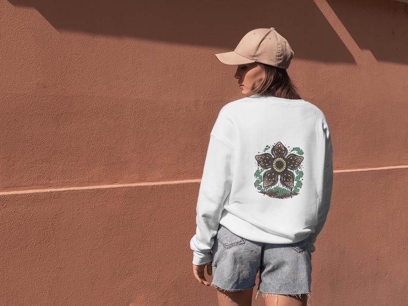 May include: White sweatshirt with a Demogorgon graphic on the back. The graphic is a detailed illustration of the creature's flower-like head, with brown and green colors. The person is wearing a tan baseball cap and denim shorts.