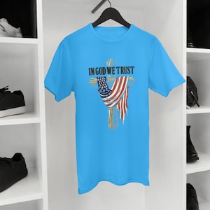 Puede incluir: Una camiseta azul claro con un dise&ntilde;o de cruz y una bandera estadounidense drapeada sobre ella. El texto "IN GOD WE TRUST" est&aacute; impreso encima de la cruz. La camiseta est&aacute; colgada de una percha negra.