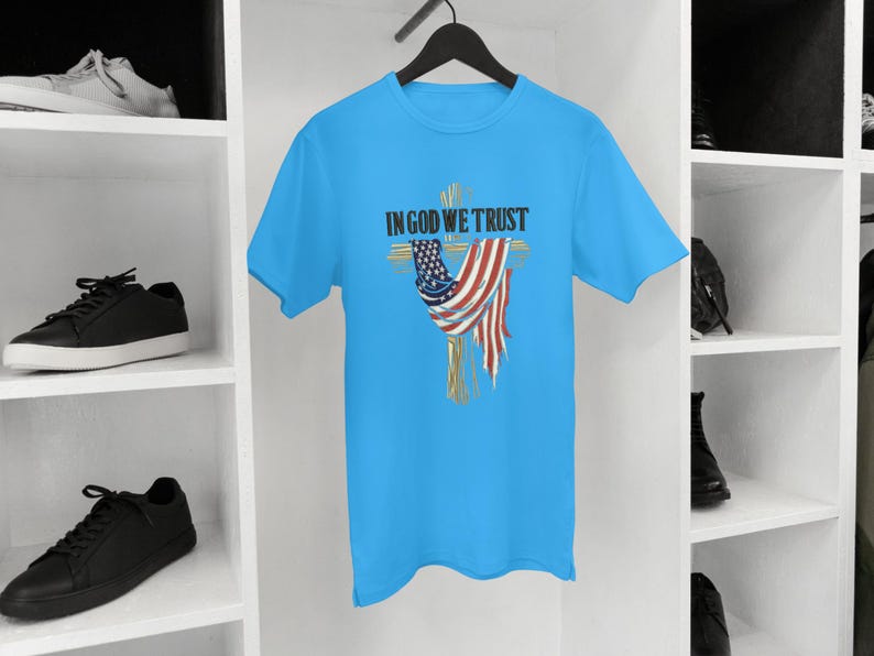 Puede incluir: Una camiseta azul claro con un dise&ntilde;o de cruz y una bandera estadounidense drapeada sobre ella. El texto "IN GOD WE TRUST" est&aacute; impreso encima de la cruz. La camiseta est&aacute; colgada de una percha negra.
