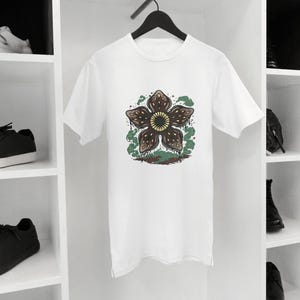 May include: White t-shirt featuring a graphic of a Demogorgon flower with brown petals, a yellow center, and green accents. The shirt is hanging on a black hanger. Black sneakers and white sneakers are on the shelves.