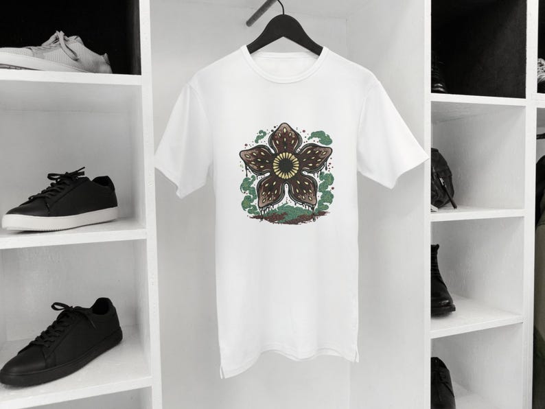 May include: White t-shirt featuring a graphic of a Demogorgon flower with brown petals, a yellow center, and green accents. The shirt is hanging on a black hanger. Black sneakers and white sneakers are on the shelves.