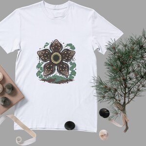 May include: White t-shirt featuring a Demogorgon graphic. The design includes a brown, flower-like creature with a yellow center, surrounded by green and brown details. The shirt is displayed with decorative items and a sprig of greenery.