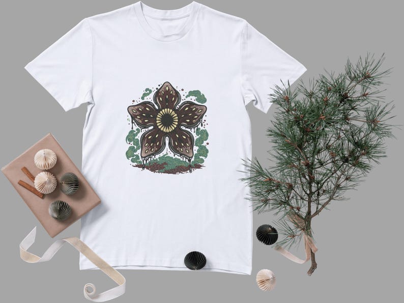 May include: White t-shirt featuring a Demogorgon graphic. The design includes a brown, flower-like creature with a yellow center, surrounded by green and brown details. The shirt is displayed with decorative items and a sprig of greenery.