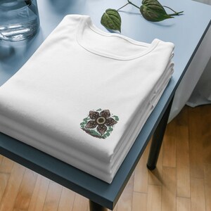 May include: A stack of white t-shirts with a crew neck. Each shirt has a Demogorgon flower graphic on the front. The shirts are neatly folded on a light blue table, with a glass vase and plant in the background.