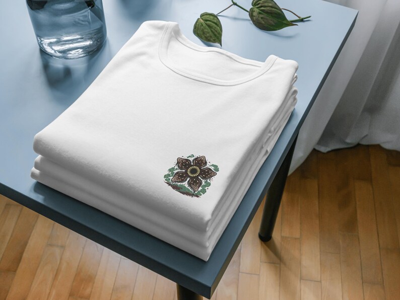 May include: A stack of white t-shirts with a crew neck. Each shirt has a Demogorgon flower graphic on the front. The shirts are neatly folded on a light blue table, with a glass vase and plant in the background.