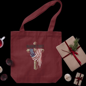Puede incluir: Bolso tote burdeos con un dise&ntilde;o de cruz con una bandera estadounidense y el texto "IN GOD WE TRUST". Un regalo envuelto con una cinta roja y vegetaci&oacute;n est&aacute; al lado de la bolsa. Tambi&eacute;n hay otras decoraciones.