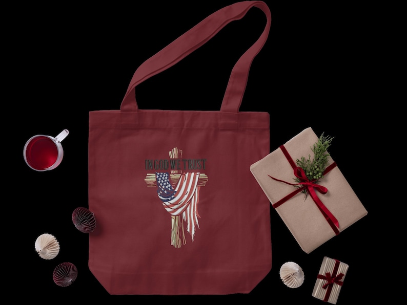 Puede incluir: Bolso tote burdeos con un dise&ntilde;o de cruz con una bandera estadounidense y el texto "IN GOD WE TRUST". Un regalo envuelto con una cinta roja y vegetaci&oacute;n est&aacute; al lado de la bolsa. Tambi&eacute;n hay otras decoraciones.