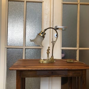 May include: A gold-toned table lamp with a white glass shade and a decorative figure on the base. The lamp is sitting on a wooden table with a drawer.