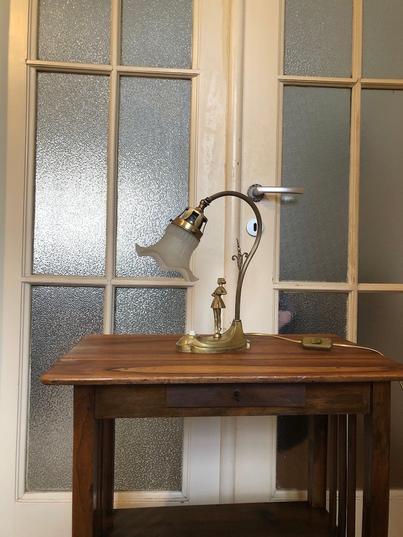 May include: A gold-toned table lamp with a white glass shade and a decorative figure on the base. The lamp is sitting on a wooden table with a drawer.
