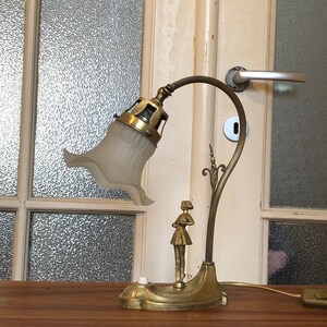 May include: A brass desk lamp with a frosted glass shade and a decorative figure of a woman. The lamp has a curved arm and a round base.