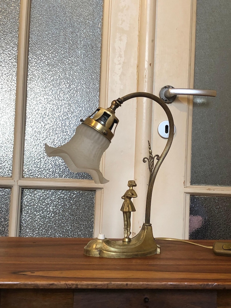 May include: A brass desk lamp with a frosted glass shade and a decorative figure of a woman. The lamp has a curved arm and a round base.