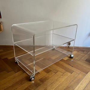 May include: A clear acrylic rolling cart with two shelves. The cart has rounded edges and sits on four small, silver-coloured wheels. The shelves are rectangular and provide storage space. The cart is designed for display or storage.