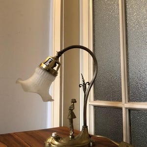 May include: A vintage brass desk lamp with a white frosted glass shade and a decorative figure of a woman. The lamp has a flexible arm and a switch on the cord.
