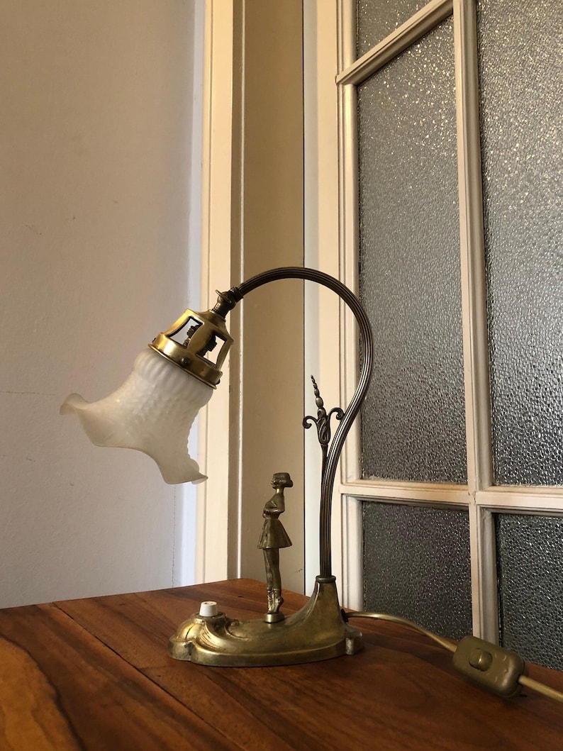 May include: A vintage brass desk lamp with a white frosted glass shade and a decorative figure of a woman. The lamp has a flexible arm and a switch on the cord.