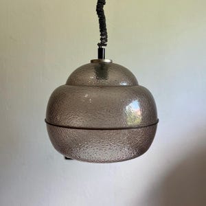 May include: A gray, textured glass pendant light with a rounded, layered design. The light fixture has a dark cord and metal accents. The glass shade has a dimpled surface and a dark metal band around the bottom.