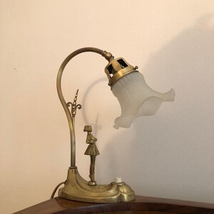 May include: A brass table lamp with a frosted glass shade and a decorative figure of a woman. The lamp has a curved arm and a base with intricate details.