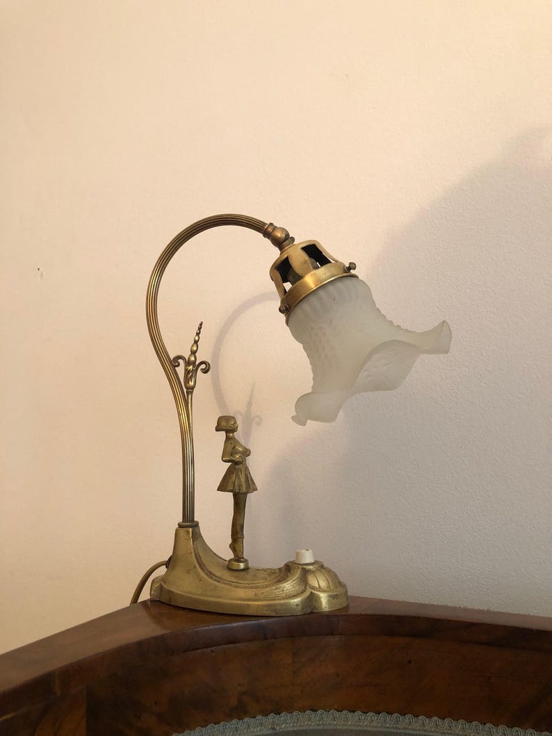 May include: A brass table lamp with a frosted glass shade and a decorative figure of a woman. The lamp has a curved arm and a base with intricate details.