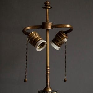 May include: A brass lamp base with two sockets and pull chains. The lamp base has a decorative design at the bottom.