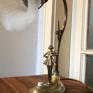 May include: A vintage brass table lamp with a frosted glass shade and a sculpted ballerina figurine. The lamp has a decorative base and a flexible arm.