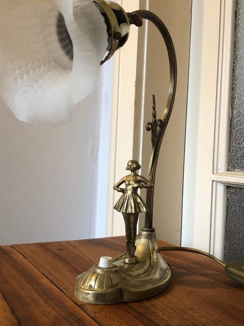 May include: A vintage brass table lamp with a frosted glass shade and a sculpted ballerina figurine. The lamp has a decorative base and a flexible arm.