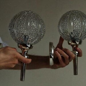 May include: Two clear glass globe sconces with a textured pattern, mounted on silver-colored metal stems and wall plates. The sconces are held by a person, showcasing their design and potential for wall lighting.