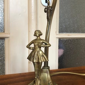 May include: A gold-toned table lamp with a ballerina figurine on top. The lamp has a decorative base and a curved arm.