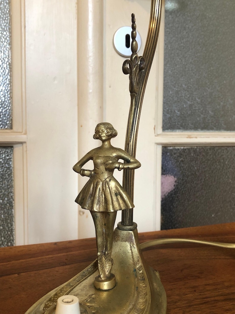 May include: A gold-toned table lamp with a ballerina figurine on top. The lamp has a decorative base and a curved arm.