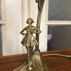 May include: A gold-toned table lamp with a ballerina figurine. The lamp has a decorative base and a curved arm.