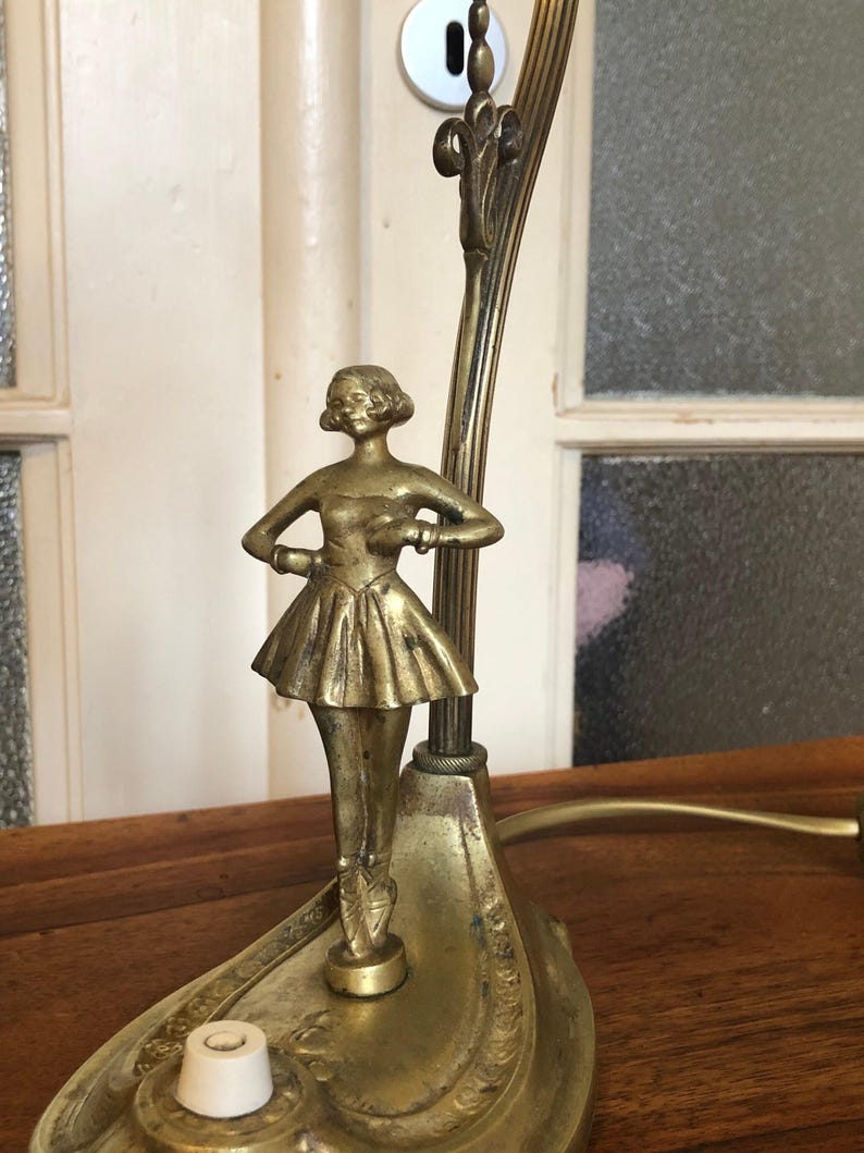 May include: A gold-toned table lamp with a ballerina figurine. The lamp has a decorative base and a curved arm.